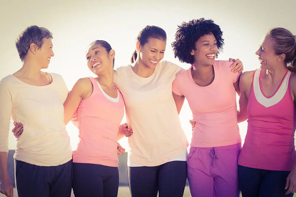 A diverse group of five women in casual athletic wear stand arm in arm, smiling and laughing together against a blurred outdoor background. They are dressed in shades of pink and white tops, suggesting a positive and supportive gathering.
